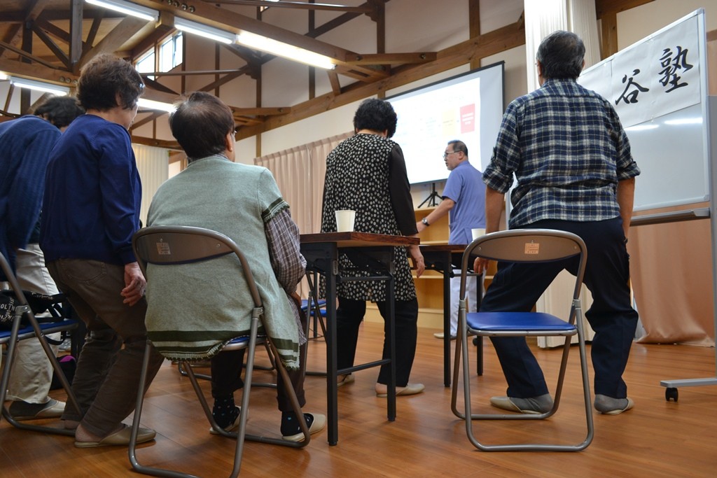 前方で中村先生が説明をする中、数人の高齢者が椅子から立ち上がりスクワット運動を実践している様子の写真