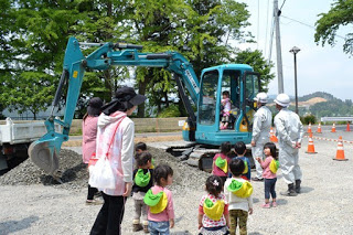 砂利の上に止まっている青いショベルカーの前に保育士に付き添われた幼児たちが数人並んでおり、作業服とヘルメットを着た男性2人がショベルカーの操縦席に乗った園児を見守っている写真