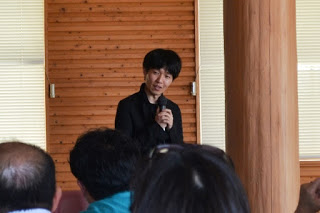 黒い服を着た男性がマイクを使って話しており、前方にはその話を聞いている複数の参加者の人々が座っている様子が写っているセミナーの写真