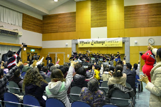 観客席からステージを見上げ、楽団の演奏に合わせて大勢の参加者が手を挙げて盛り上がっている音楽交流会の写真