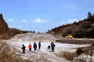 周囲を木々に囲まれた造成地のような場所で、奥は土が盛られ、黄色いシャベルカーがあり、数名の人たちが調査に訪れている様子を写した写真