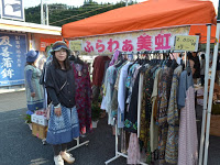 オレンジ色のテントの下に「ふらわぁ美虹」と書かれた看板が掲げられ、その前にたくさんの服が掛けられたラックが並び、店の前に立った女性がカメラの方を向いている写真