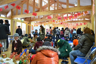 木のぬくもりが感じられる建物の中で、多くの参加者が床に座り、話したり、ブロックなどで子どもたちが遊んでいる様子が写っている室内の写真