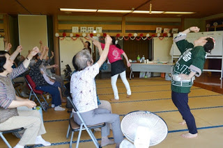 畳の上で前方に立っている3人の女性が片手を上げている動きをし、椅子に座った参加者たちが、片手を大きく上げて元気に体操をしている様子が写っている写真