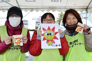 「南三陸町ボランティアセンター」と書かれたベストを着た女性3人が、キャラクター「にこま～る」のイラストが描かれたマスコットなどのグッズを持って笑顔で並んでいる写真