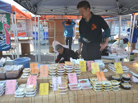 テントの下に置かれた台の上に様々な種類のパック詰めされた食品がたくさん並べられ、後ろに店員の男性が写っている写真