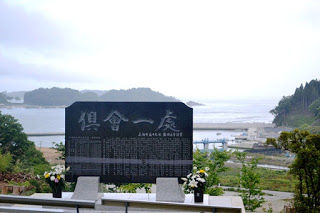 「倶會一處」の文字とたくさんの名前が刻まれた黒い石碑が、漁港と海を見下ろす高台に建っている写真