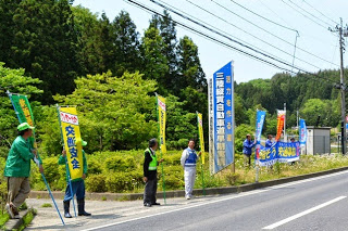 数人の男性が道路脇に立ち、それぞれが交通安全を呼びかける旗を持って通行人や車に向けて注意喚起をしている様子が写っている交通安全運動の写真