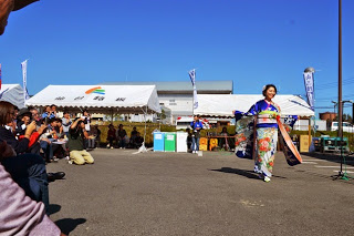 青色で花柄の華やかな着物を着た女性が、広場の中央を歩きながら観客に笑顔を向けている写真