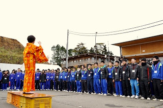 オレンジ色の着物を着た人が仮設の台の上に立って指揮をとり、大勢の中学生たちが並んで歌っている様子の写真