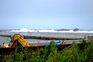 防波堤の外で波が高くうねり、白波が続き手前には工事用の黄色いショベルカーが壁際に置かれている写真