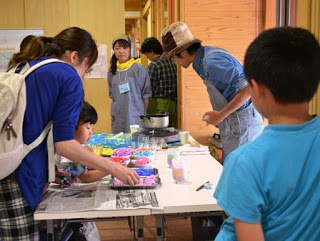 複数の人々が室内でテーブルを囲み、それぞれが手元の作業に集中して取り組んでいる様子が写っている写真