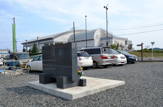 数台の車が停まった砂利の駐車場に、黒い石碑が立っている写真