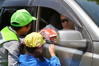 緑色の帽子をかぶった男性と青いスモッグを着て黄色の帽子をかぶっ女の子が車の窓越しに乗っている人とやり取りをし、交通安全のお守りなどが入った袋を手渡ししている写真