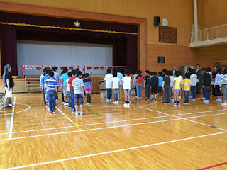 先生や大人たちが見守る中、体育館のステージ前に児童が整列している写真