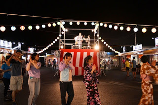 夜の祭りの会場で、紅白幕が張られたやぐらを囲んで浴衣姿の人々などが盆踊りを踊っている様子を写した写真