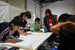 生徒が立ったり座ったりしながら模造紙にペンで何かを書き込んでいて、先生が笑顔で見守っている様子が写っている写真