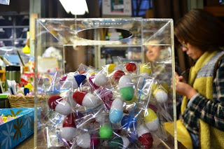 奥には2人の女性が立つ店内に置かれた透明な抽選箱の中に、カラフルなカプセルが沢山入っている写真