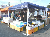 青いテントの下に白い発泡スチロールの箱や黄色・オレンジ色のプラスチックケースが整然と並べられた屋台の中に黒いエプロンをつけた男性が2人並んで立っている写真