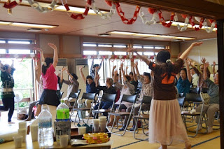 講師の指導に合わせて、畳の部屋で多くの参加者が椅子に座ったまま両手を上に伸ばして体操をしている様子が写っている写真