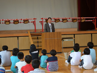 和太鼓が飾られた舞台の前で、スーツ姿の校長先生が演台に立ち、体育座りした子どもたちに話している写真