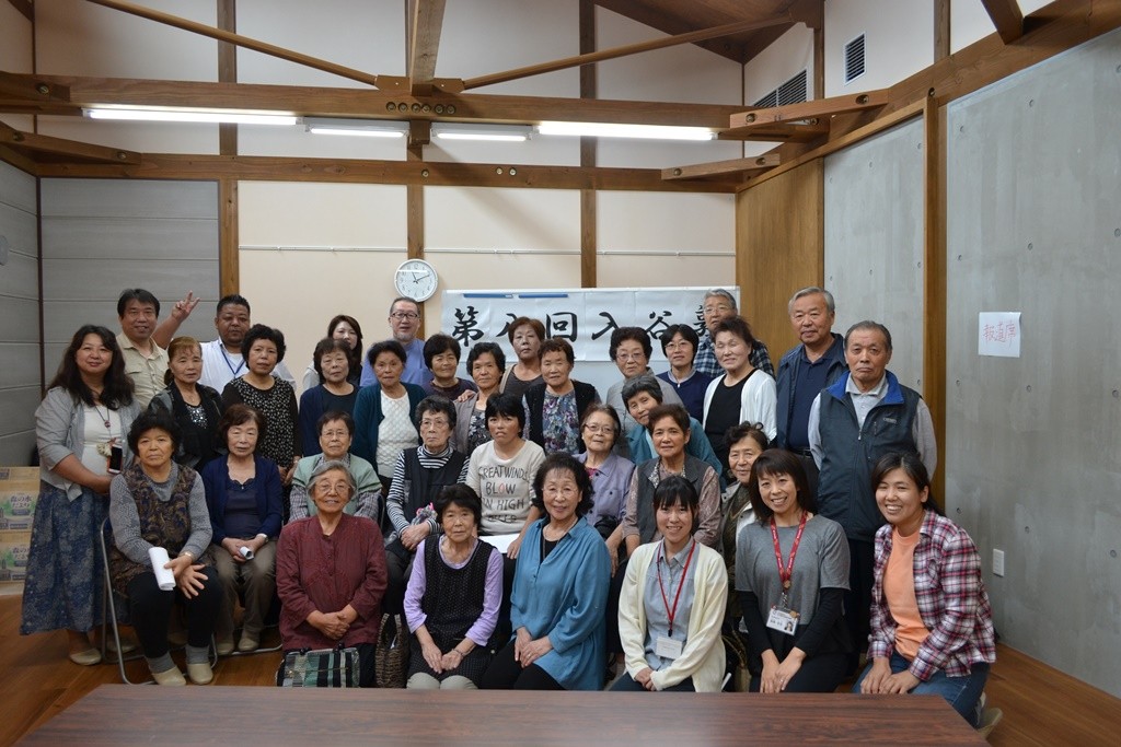 木造の部屋で『第八回入谷塾』と書かれた紙が貼られたホワイトボードの前で参加者が並んだ集合写真