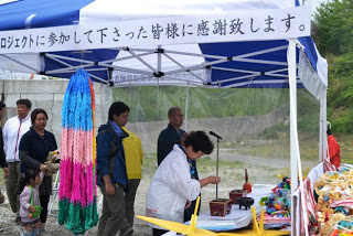 「プロジェクトに参加して下さった皆様に感謝致します」と書かれたテントの下で、白いシャツの女性がお焼香をあげており、後ろにもお焼香の列ができている様子の写真