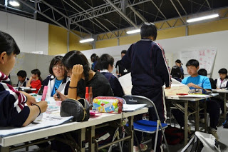 生徒たちが複数のグループに分かれて机に向かい、集中して話し合いをしている様子が写っている写真