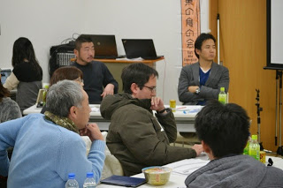 正面にスクリーンがある会議室で、話を聞いている男性たちの後ろ姿と、発言者の方向を見つめる二人の男性の表情が印象的な写真