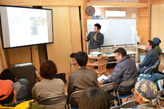 前方のスクリーンに資料を映し説明する講師と椅子に座って熱心に聞いている参加者たちの様子の写真