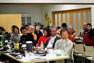 参加者たちが円卓に座り、食事や飲み物を楽しみながらスピーカーの方に注目している様子を写した写真