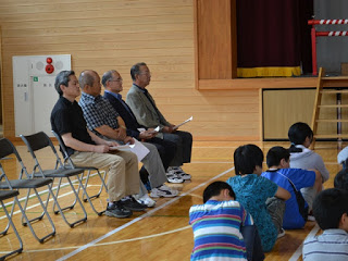 体育館の左端に並んだ椅子に座る4人の男性講師が、舞台の方向を見て話を聞いている写真