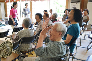 高齢の女性たちが椅子に座ったまま手を挙げたり笑ったりしながら、和やかにレクリエーションに参加している様子が写っている写真