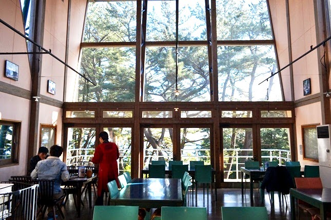 天井まで届く大きなガラス窓から松林と自然光が差し込む室内で、2人の客がテーブルに座り、赤い服を着た女性が接客をしている様子を写した写真