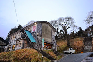 傍に軽トラックが止まっている坂道を上がった先にある木造二階建ての建物が、夕方の空の下に佇んでいる写真