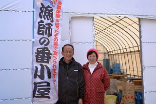 白いテントの入り口で「漁師の番小屋」と書かれた旗の横に立ち、笑顔で並んでいる村岡さん夫婦を写した写真