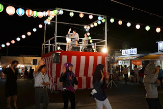 色とりどりの提灯が飾られた夜の祭り会場で、やぐらに上った人々が踊っており、その周りを囲んで一列に並んだ来場者たちが、一緒に盆踊りをしている様子の写真