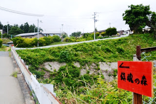 舗装された歩道の脇に「鎮魂の森」と書かれた赤い案内板が立てられ、奥には緑の丘と住宅が見える風景が広がっている写真