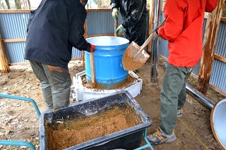 青いドラム缶を中心に、3人の作業者がスコップで周囲に土や籾殻を詰めて断熱施工をしている様子を写した、炭窯の設置工程の写真