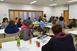 木製の引き戸が並ぶ部屋で、複数のテーブルに分かれて着席した参加者たちが講師の話に耳を傾けている写真