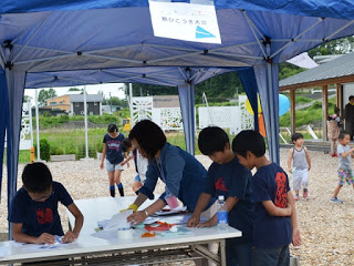 青いテントの下で男の子3名が机の上で紙飛行機を作っている様子を写した写真
