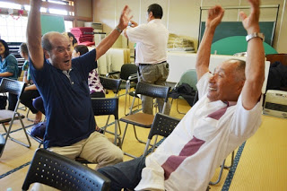 高齢の男性2人が椅子に座りながら両手を上にあげて笑顔を見せ、他の参加者たちも周囲で楽しそうに過ごしている様子が写っている写真