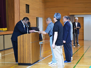 演台の前で4人の男性講師が一列に並び、向かい合って立つ校長先生から賞状の様な物をチェック柄のシャツを着た男性が代表で受け取っている写真