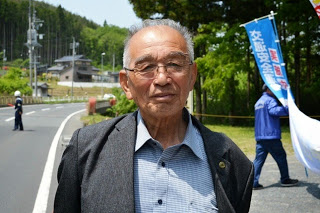 道路わきに立つ、背広姿でメガネをかけた菅原塚夫さんを正面から写した写真