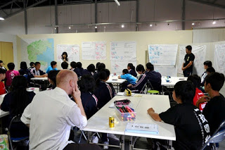 壁に貼られた模造紙の前で女性が話し、複数の生徒が前を向いて座って話を聞いている様子が写っている写真