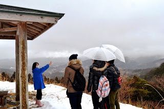 雪が積もる展望所でガイドの女性が山々を指さして説明し、数人の来訪者が傘をさして熱心に話を聞いている様子の写真