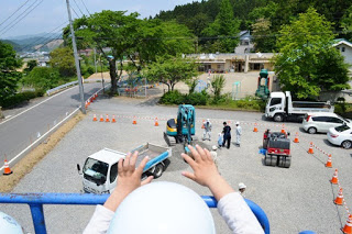 バケットから見下ろした、地上の工事車両や保育所の建物、遠くの町などの景色をヘルメットをかぶった園児の頭越しに撮影した写真
