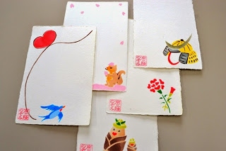 左下に「南三陸のぞみ」と書かれた印鑑が押されたハガキに、黄色の兜や、桜の花びらが舞い降りてくるリス、赤いカーネーション、タケノコの親子、ハートマークとツバメが飛んでいる様子を描いた絵葉書が重ねて置かれている写真