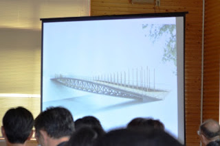 水上にかかる橋のデザイン画がスクリーンに映されていて、参加者たちがそれを見ている講演会の様子の写真