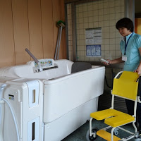 介護用の特殊な浴槽のそばに女性が立ってリモコンを持っており、浴槽の横には車輪付きの黄色い入浴用椅子が置かれている写真